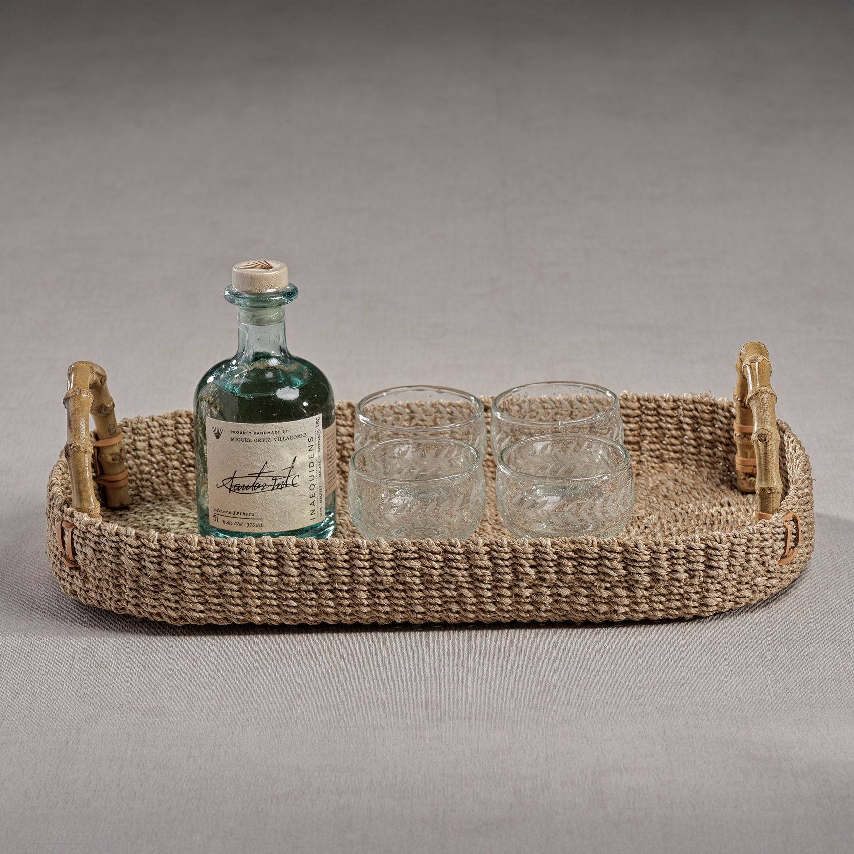 The Zodax Bamboo Handle Abaca Tray Collection displays a glass bottle with a cork and three glass tumblers, all elegantly arranged on a neutral surface.
