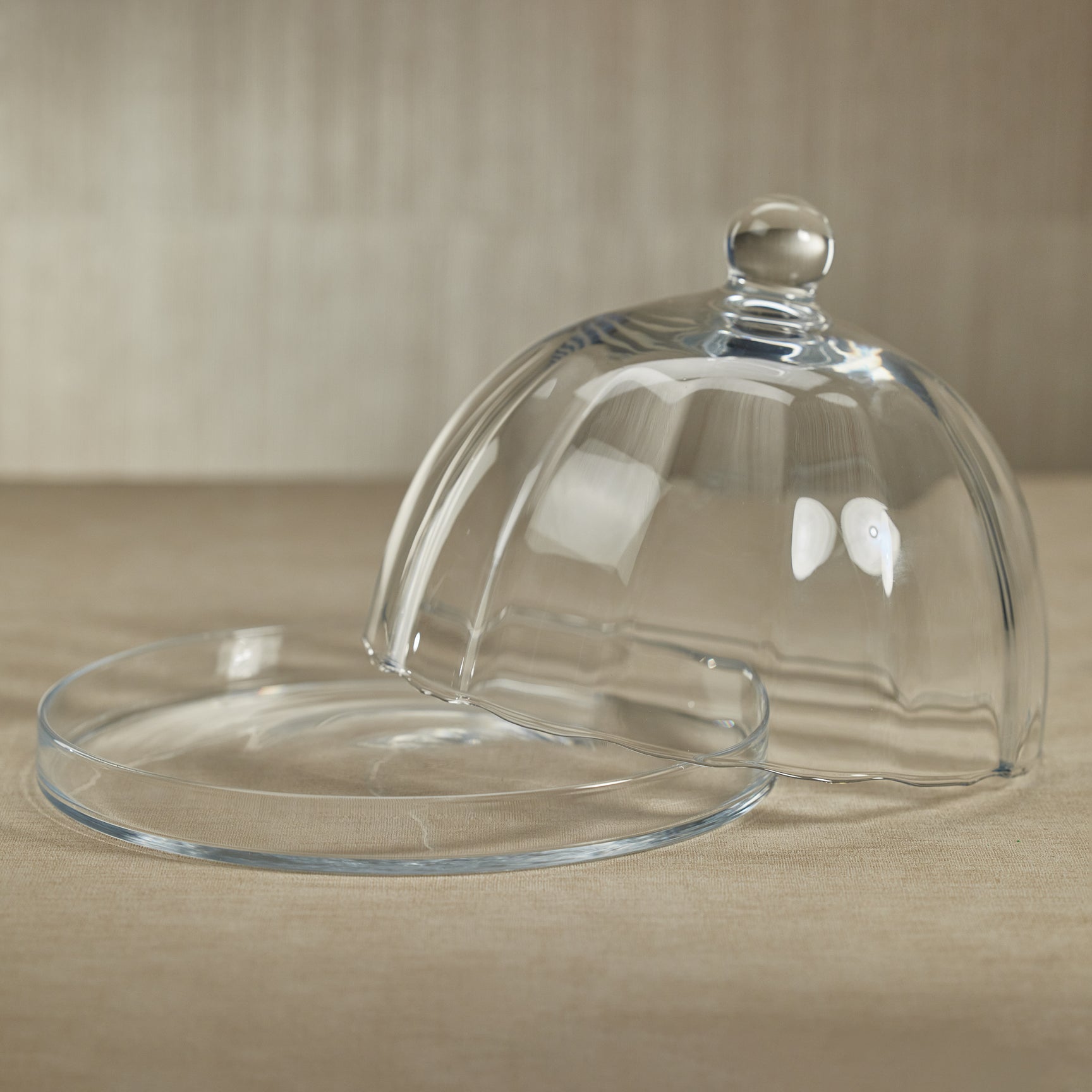 The Zodax Loulou Optic Pastry Glass Plate with Cloche features a clear glass cloche tilted off its matching round plate, both elegantly displayed on a beige surface.