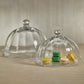 Two Zodax Loulou Optic Pastry Glass Plates with Cloches are displayed: one dome is empty, the other covers five assorted yellow, green, and beige macarons on a beige surface—creating an elegant dessert presentation.