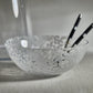The Zodax Liguria Confetti Bowl, made of patterned glass with a white dappled design, rests on a light surface; inside lie two black chopsticks with white tips—an elegant accent for any setting.