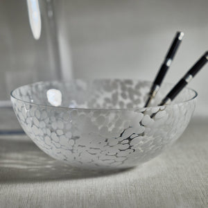 The Zodax Liguria Confetti Bowl, made of patterned glass with a white dappled design, rests on a light surface; inside lie two black chopsticks with white tips—an elegant accent for any setting.