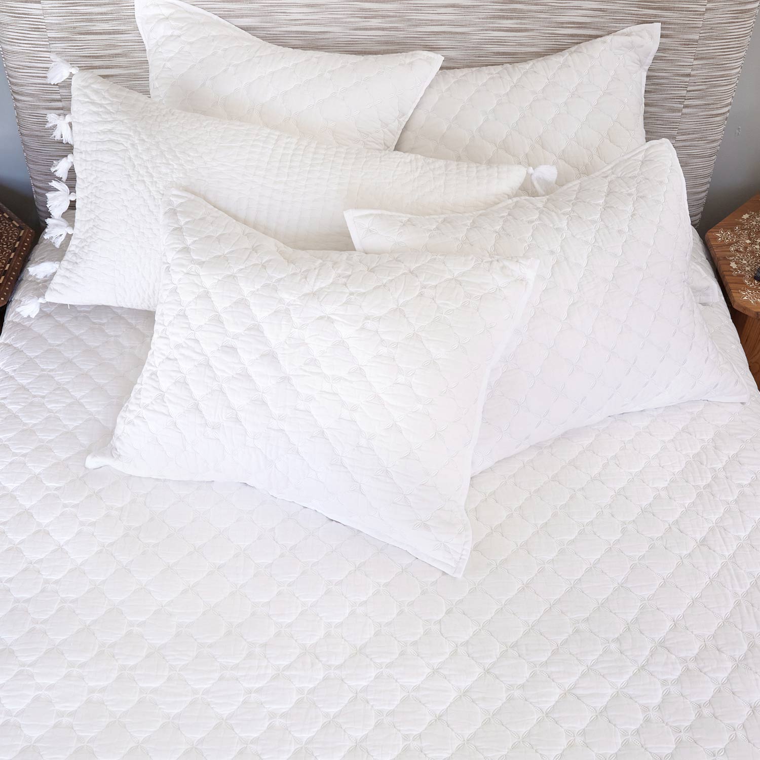 Five pillows from the John Robshaw Layla Bedding Collection, their white quilted texture set against a pristine white cotton voile quilt bedspread, are artfully arranged with a textured headboard and flanked by two wooden nightstands—evoking the tranquil elegance and inviting oasis atmosphere inspired by Mughal gardens.