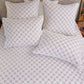 A neatly made bed with the John Robshaw Layla Lavender Coverlet and five matching pillows, all showcasing a subtle gray geometric pattern on white cotton voile.