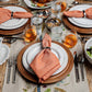 A table set for four with white plates, wooden chargers, peach napkins in rings, Juliska’s Graham 5 Piece Flatware Set (polished), glasses of water and amber drinks, plus salad and snacks on a beige floral tablecloth—perfect for casual entertaining.
