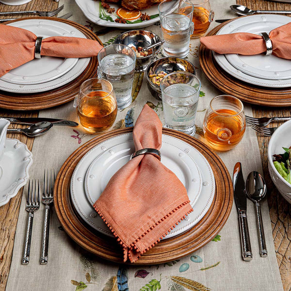 A table set for four with white plates, wooden chargers, peach napkins in rings, Juliska’s Graham 5 Piece Flatware Set (polished), glasses of water and amber drinks, plus salad and snacks on a beige floral tablecloth—perfect for casual entertaining.
