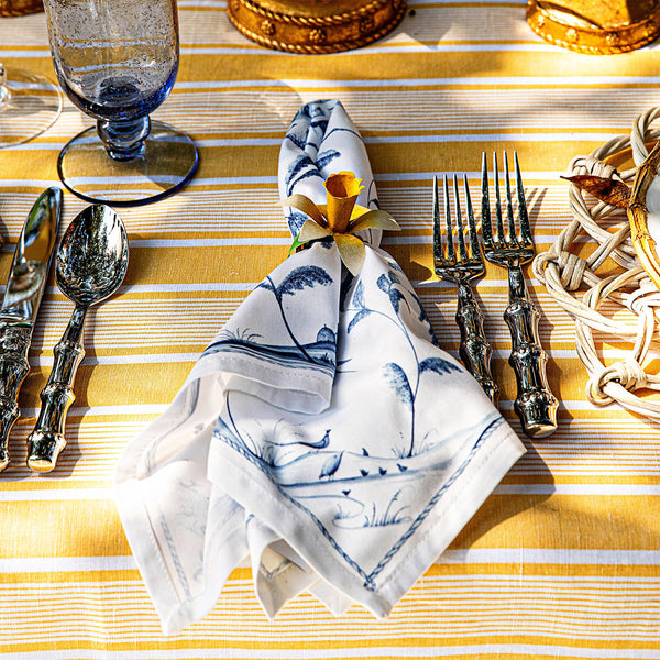 A place setting on a yellow striped tablecloth showcases Juliska’s Meadow Walk Daffodil Napkin Ring Set, three forks, a knife, spoon, glass tumbler, and napkin in a ring—an inviting touch for elegant dining.
