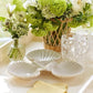 The Aerin Shell 3-Bowl Dish features three white, scallop-shaped bowls, displayed with three clear spotted glasses, a small vase of white flowers, and a wicker basket filled with green and white blooms.
