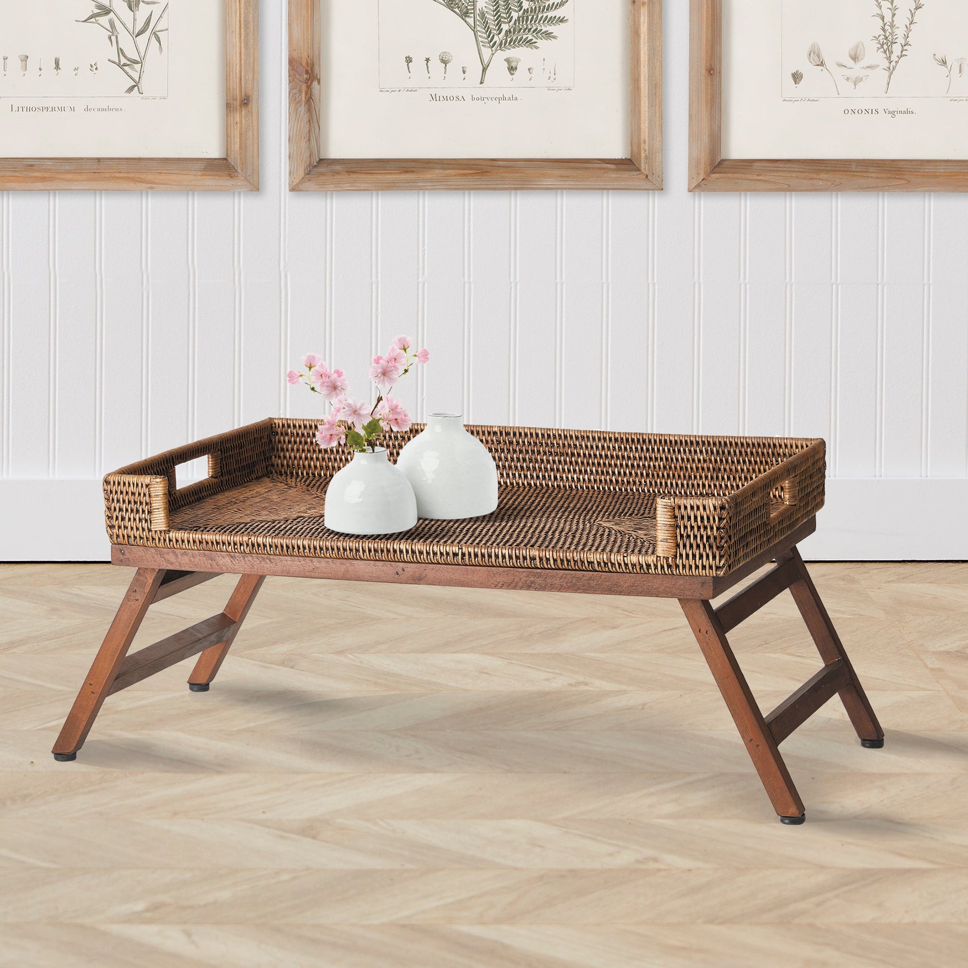 The Burma Rattan Lap Tray Table by Napa Home & Garden, shown with two white vases and pink flowers, features folding legs for easy laptop use or breakfast in bed and is set on a wooden floor with framed botanical prints behind.