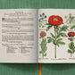 An open book from Taschen titled "Basilius Besler" rests on a green surface, showing botanical illustrations of peonies and blue flowers with Latin text; an orange ribbon bookmark is visible.
