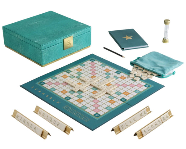 Introducing the Scrabble Del Mar Shagreen Edition by WS Game Company, a premium set featuring luxurious textures with deluxe components: a game board, letter tiles, tile racks embossed with words, a sand timer, a notebook, and an elegant teal storage box adorned with faux-shagreen on a marble table.