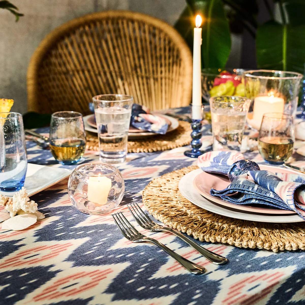 A Juliska Ikat Tablecloth with hand-stitched trim sets the base for woven placemats, patterned napkins, plates, and glassware. Lit candles, a wicker chair, and green plants add charm against a sun-faded blush backdrop.