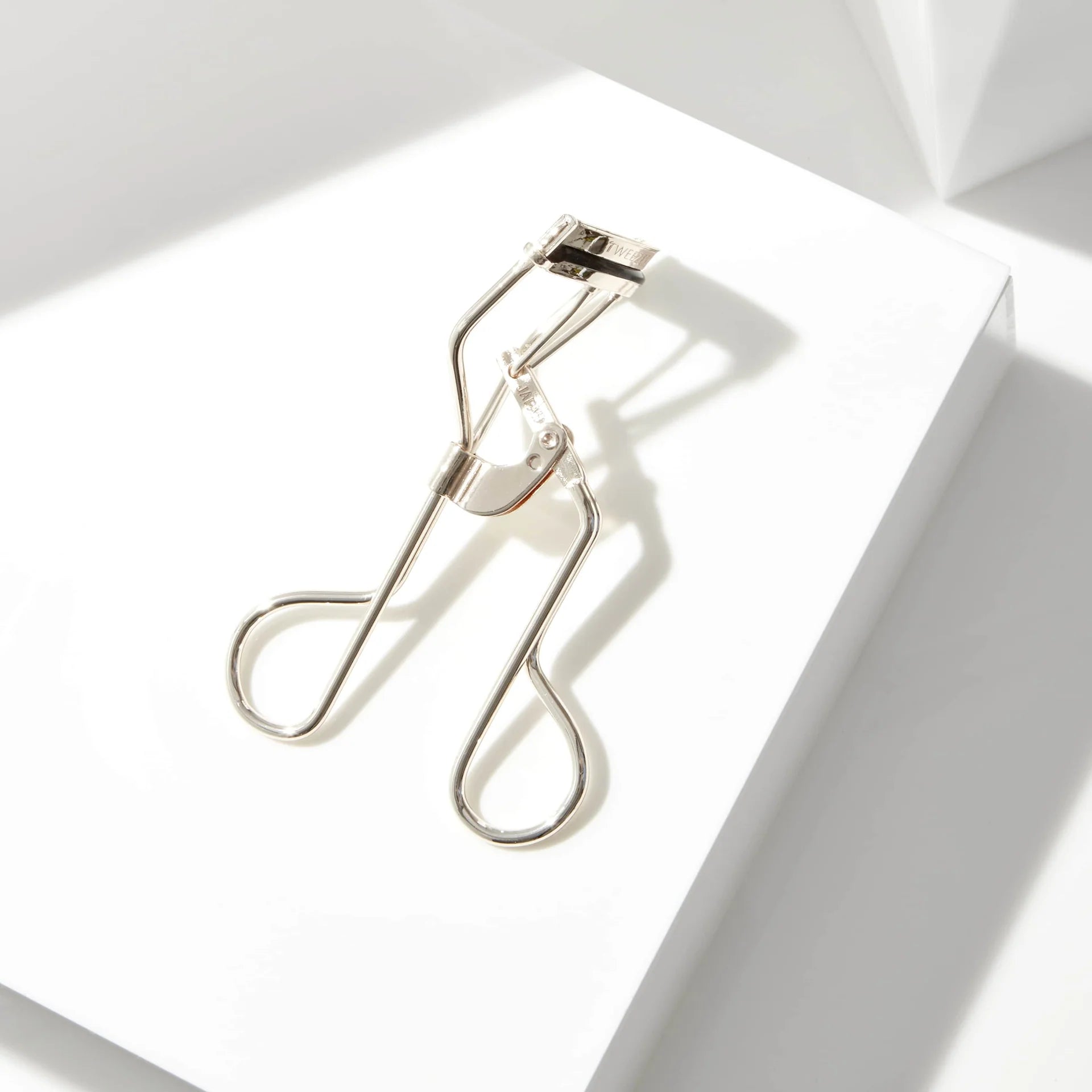 The Tweezerman Curl 38 Eyelash Curler rests on a white surface, its sleek design casting shadows—ideal for enhancing almond-shaped eyes with a natural lift.