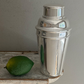 The Hôtel Silver Vintage Cocktail Shaker with Built-in Citrus Juicer rests on a wooden surface beside a whole lime, set against a plain gray wall.