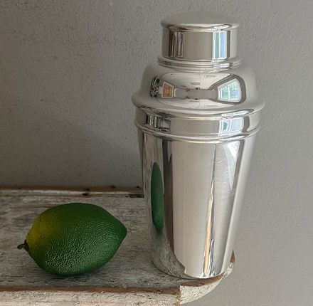The Hôtel Silver Vintage Cocktail Shaker with Built-in Citrus Juicer rests on a wooden surface beside a whole lime, set against a plain gray wall.
