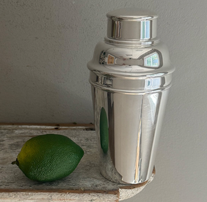 The Hôtel Silver Vintage Cocktail Shaker with Built-in Citrus Juicer rests on a wooden surface beside a whole lime, set against a plain gray wall.
