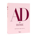 A pink hardcover book titled “AD At Home: Architectural Digest,” published by Penguin Random House, features large red letters on the cover and offers interior design inspiration while celebrating unique homes.