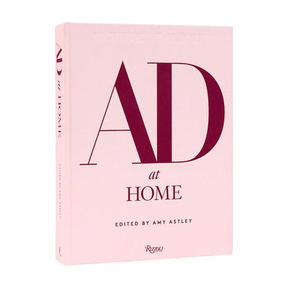 A pink hardcover book titled “AD At Home: Architectural Digest,” published by Penguin Random House, features large red letters on the cover and offers interior design inspiration while celebrating unique homes.