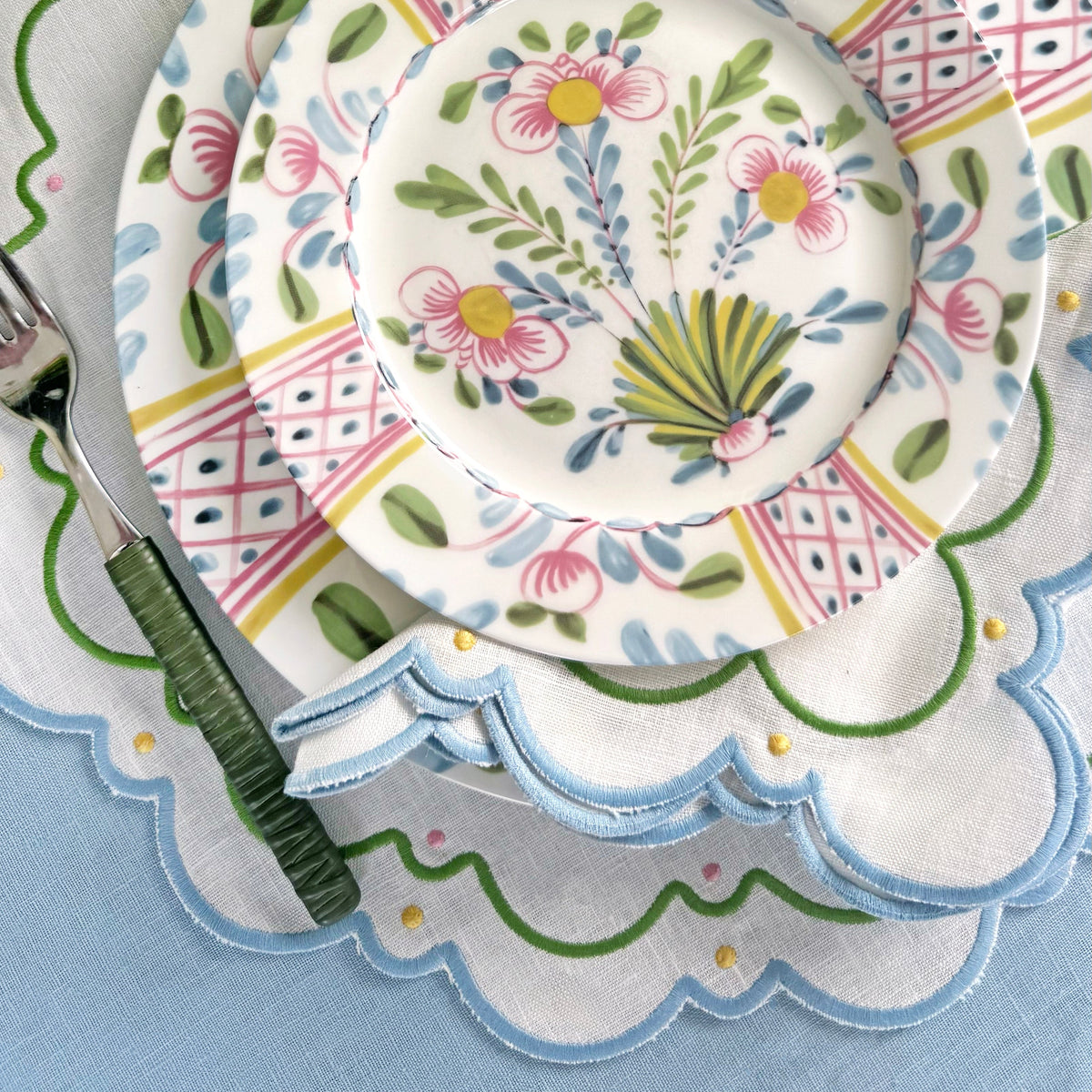 A stack of floral patterned plates from the Sugus Table Linen Collection set on scalloped embroidered placemats with a green-handled fork.
