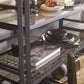 The Tom-Tom Etagere by Century Furniture displays stacked dark dishes, books, decorative objects, and bowls. In the foreground, a wooden chair and nesting tables are partially visible.