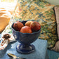 A blue pedestal bowl from the Juliska Le Panier Mystic Knot Denim Dinnerware and Serveware Collection, filled with five pomegranates, sits atop a large charger on a blue tablecloth next to a spoon and napkin, with floral pillows in the background.