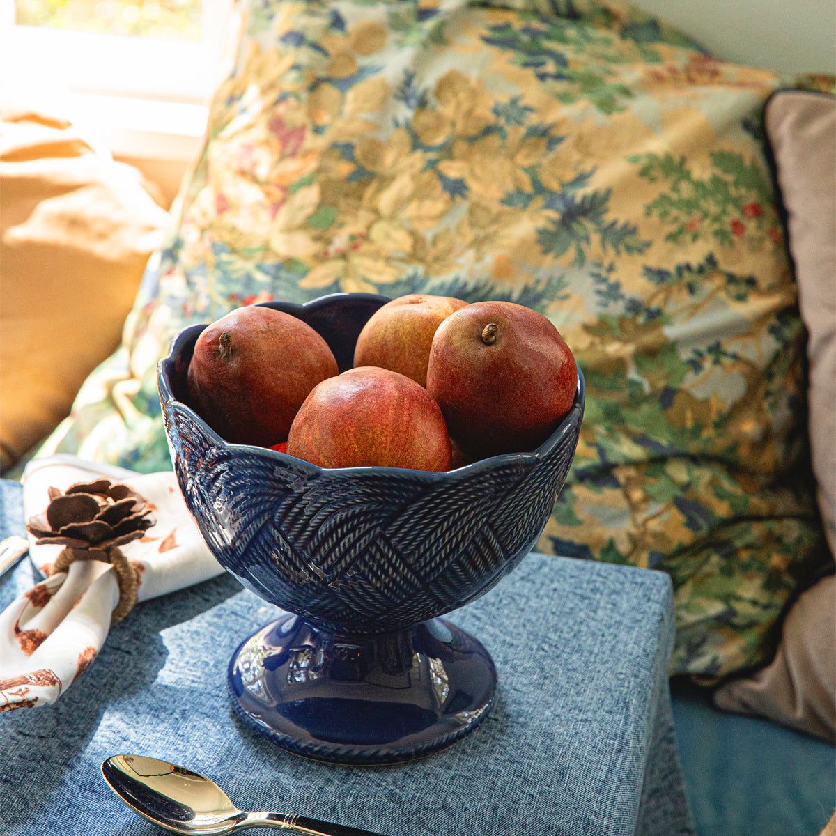 A blue pedestal bowl from the Juliska Le Panier Mystic Knot Denim Dinnerware and Serveware Collection, filled with five pomegranates, sits atop a large charger on a blue tablecloth next to a spoon and napkin, with floral pillows in the background.