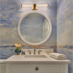 The bathroom features the Albert Kabibe Shell Mirror by Made Goods with a polished shell frame, wall-mounted light, white countertop, sink, faucet, vase of flowers, soap, and folded towel set against blue patterned wallpaper.