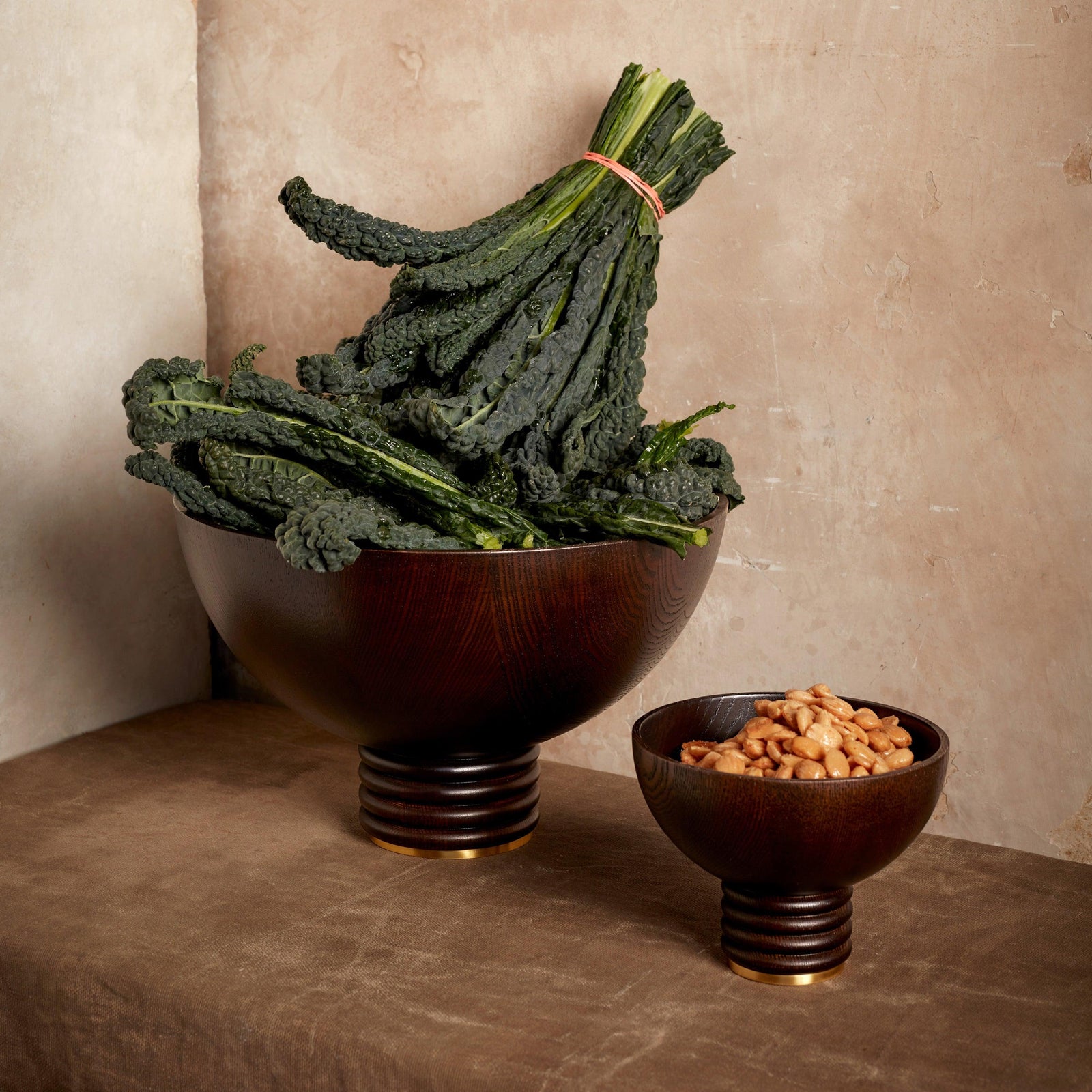 A large Alhambra Bowl by L’Objet filled with kale sits next to a smaller Alhambra bowl of peanuts, both serving as decorative accents on a brown surface against a beige wall.
