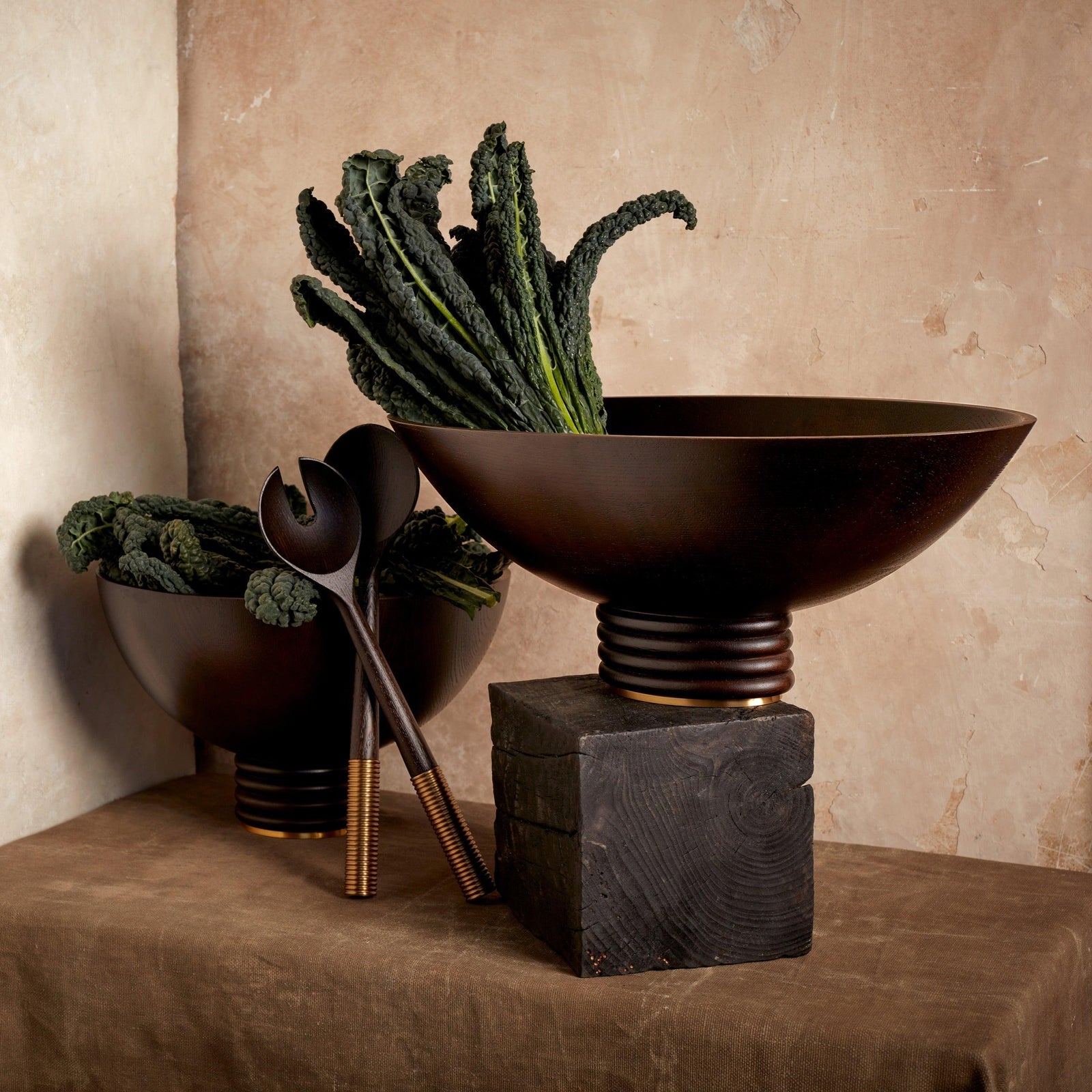 Two dark bowls from the Alhambra Bowl Collection by L’Objet, filled with kale and paired with wooden utensils and a black block, accent a brown tablecloth—recalling the charm of Alhambra Gardens.