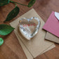 The Simon Pearce Highgate Heart "loved" glass paperweight rests on brown envelopes by a pink envelope and green plant on a wooden table, making it a thoughtful gift.