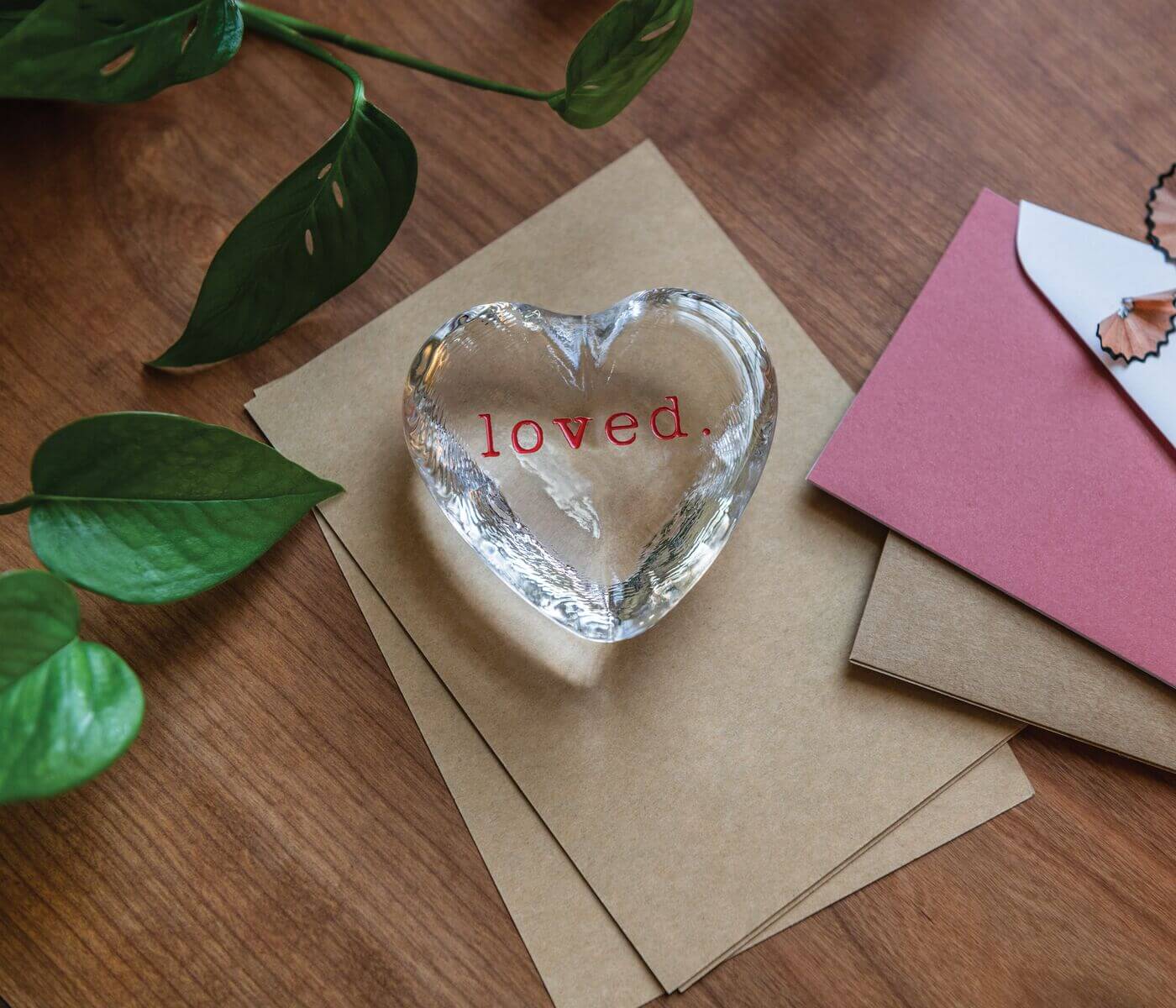 The Simon Pearce Highgate Heart "loved" glass paperweight rests on brown envelopes by a pink envelope and green plant on a wooden table, making it a thoughtful gift.