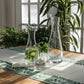 Three Simon Pearce Barre Carafe Collection pieces, one filled with water, cucumber, and mint, rest on a wooden table with a leafy runner in a bright room featuring greenery and large windows.