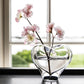 The Romance Large Vase by Simon Pearce—crafted from lead-free glass in a clear, heart-shaped design—holds two pale pink flower branches, displayed on a dark surface before a window with a softly blurred outdoor view.