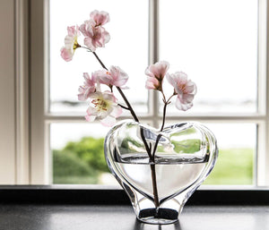 A large Simon Pearce Romance Vase, hand-sculpted with pink flowers, rests elegantly on a black surface against a window background.