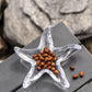The Starfish Glassware Collection by Simon Pearce features a clear star-shaped bowl holding small brown seeds, set on a gray surface with rocks—perfect as a beach-inspired décor accent or a unique summer host gift.