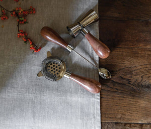 The Simon Pearce Acacia Bar Tool Set—featuring a double-sided jigger, bar spoon, and julep strainer with wooden handles—is displayed on a gray cloth beside orange berries atop a wooden table.