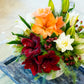 The Live Amaryllis Holiday Floral Arrangement by Hive Floral Studio features orange, red, and white flowers—including white rose lilies—greenery, pine branches, and red berries in a white vase on a blue patterned tray.