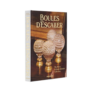 A hardcover book from Assouline, "Boules D'Escalier: The Flávia & Frank Abubakir Collection," features four ornate glass and brass staircase finials on its cover.