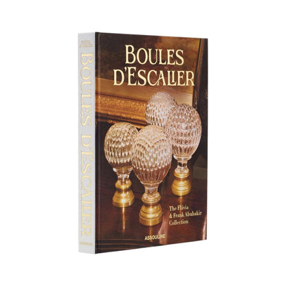 A hardcover book from Assouline, "Boules D'Escalier: The Flávia & Frank Abubakir Collection," features four ornate glass and brass staircase finials on its cover.