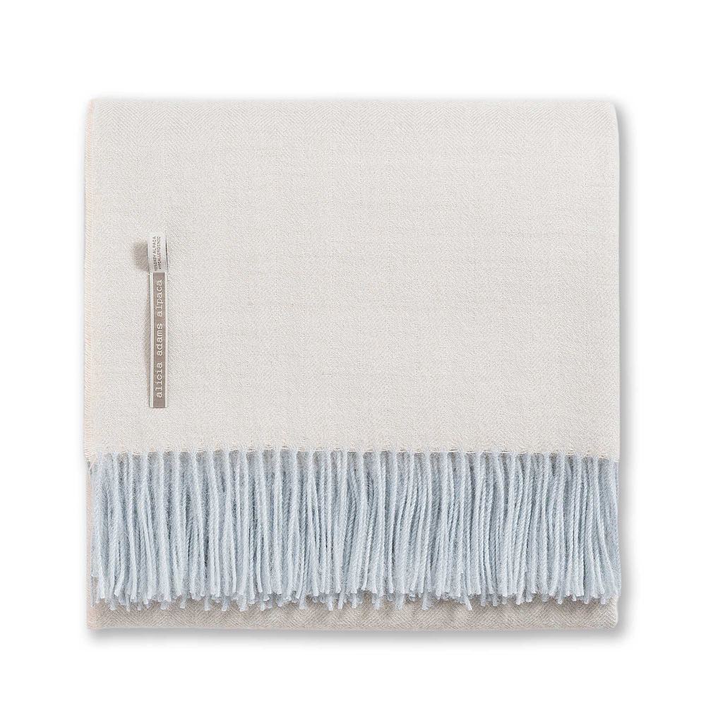 The Classic Herringbone Throw Collection by Alicia Adams Alpaca features a folded cream alpaca blanket with a tag and light blue fringe, fair-trade crafted in Peru, shown on a white background.