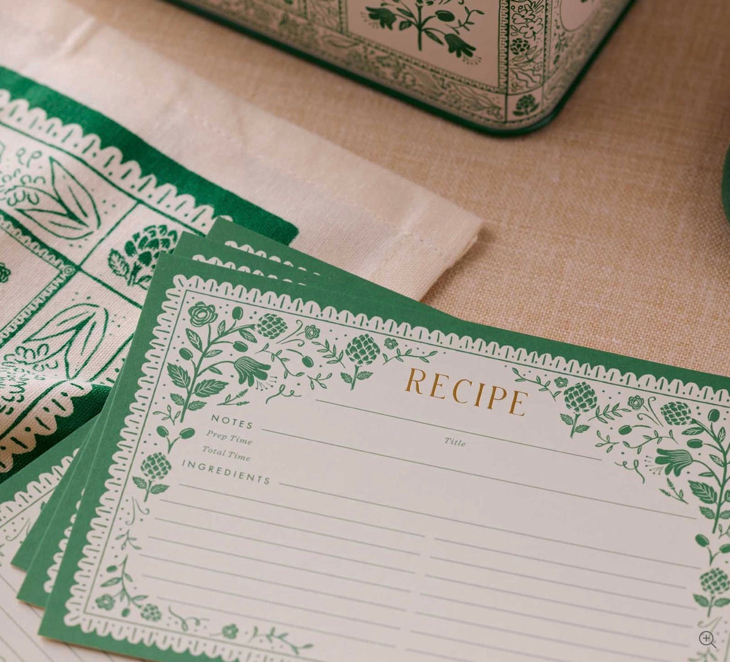 A close-up of Rifle Paper Co. Artichoke Recipe Cards with green floral borders, displayed on a beige tablecloth beside a patterned cloth and a decorative tin.
