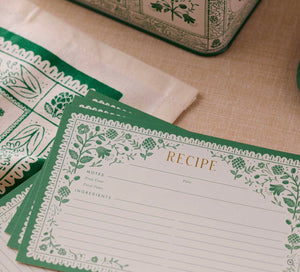 A close-up of Rifle Paper Co. Artichoke Recipe Cards with green floral borders, displayed on a beige tablecloth beside a patterned cloth and a decorative tin.