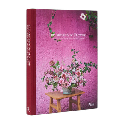 The Rizzoli book “The Artistry of Flowers” stands upright against a pink wall, its cover showcasing a floral arrangement atop a wooden table—an inspiring homage to the creativity of floral designers and indoor plant enthusiasts.