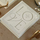 The PrintWorks "A Story of Love Wedding Album" in white rests on a beige surface with metallic decor and a plant—ideal for preserving your cherished wedding memories.