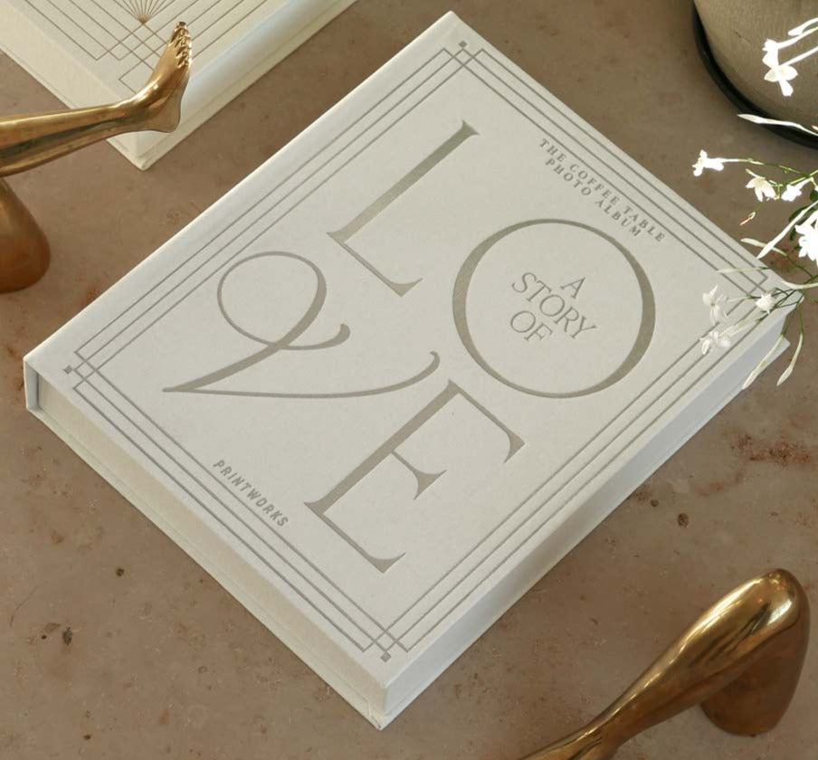 The PrintWorks "A Story of Love Wedding Album" in white rests on a beige surface with metallic decor and a plant—ideal for preserving your cherished wedding memories.