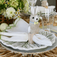 A folded napkin is displayed in a Kim Seybert Easter Bunny Napkin Ring on a white ceramic plate with a leaf design; glassware and flowers add a charming touch in the background, creating festive spring décor.