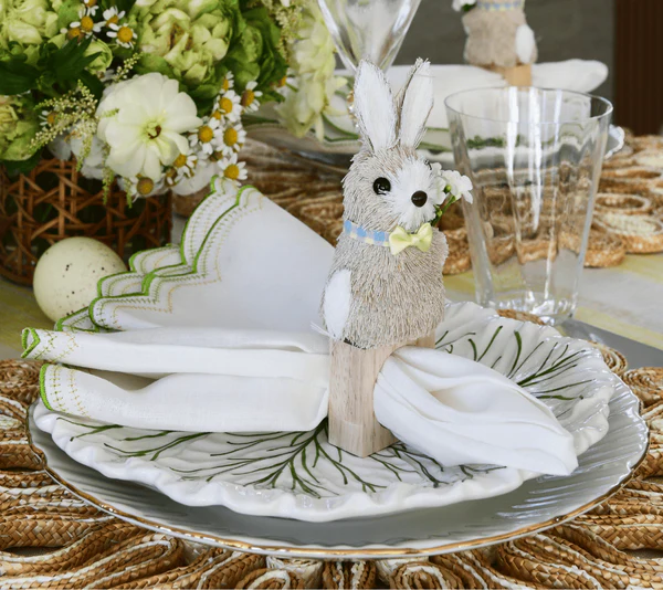 A folded napkin is displayed in a Kim Seybert Easter Bunny Napkin Ring on a white ceramic plate with a leaf design; glassware and flowers add a charming touch in the background, creating festive spring décor.