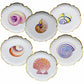 The Shell Bread and Butter Plate Collection by Anna Weatherley features six ornate porcelain plates with gold trim, each showcasing a unique hand painted shell at the center and arranged in a circular design.