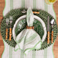 A place setting from Juliska’s Cabana Stripe Tabletop Collection features green and white plates, bamboo-handled cutlery, a striped green napkin, and a decorative placemat atop a cabana stripes table runner.