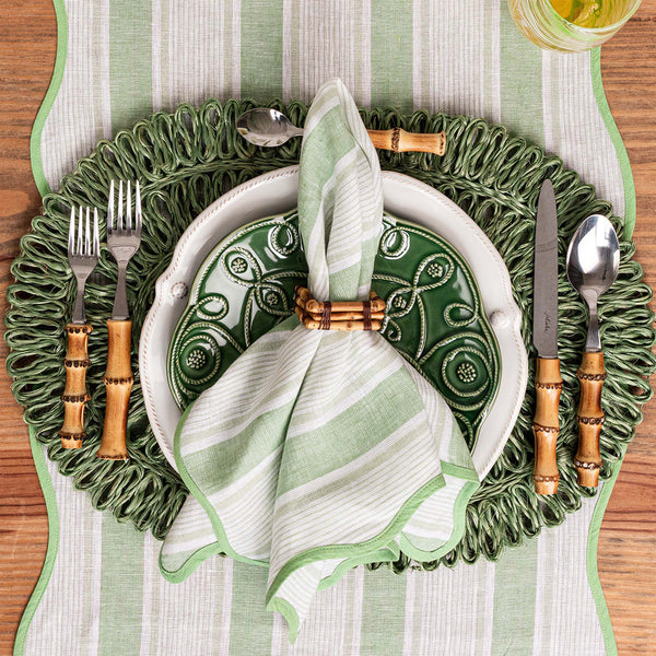 A place setting from Juliska’s Cabana Stripe Tabletop Collection features green and white plates, bamboo-handled cutlery, a striped green napkin, and a decorative placemat atop a cabana stripes table runner.