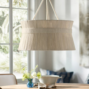 The Cassidy Chandelier by PALECEK, a beige drum-shaped fixture crafted from abaca rope, hangs above the dining table with floral accents and a bowl; natural light streams in through large windows. Professional installation recommended.